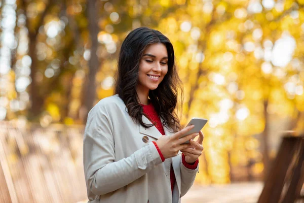 Hermosa joven dama en el mensaje de lectura traje de otoño en el teléfono inteligente en el hermoso parque —  Fotos de Stock