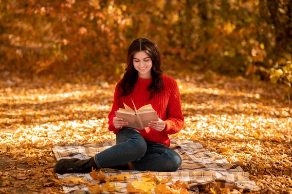 Piękna młoda dama w czerwonym swetrze siedzi na kocu piknikowym w jesiennym parku i czyta ciekawą książkę — Zdjęcie stockowe