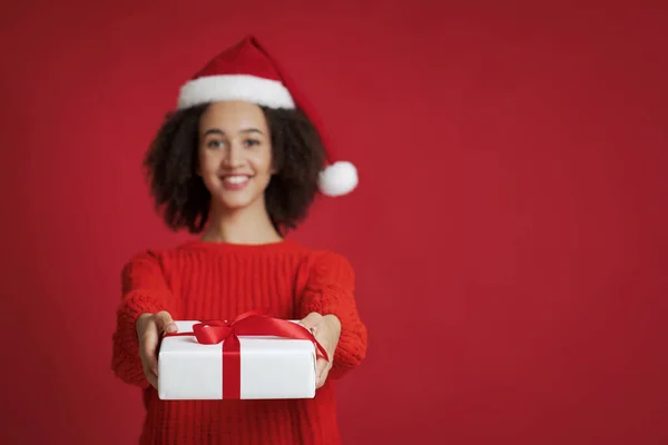 Alegre joven afroamericano hembra en Santa Claus sombrero y suéter da regalo y mira a la cámara —  Fotos de Stock