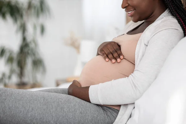 Zwarte zwangere vrouw zit op bed bij het omhelzen van de buik, zijaanzicht — Stockfoto