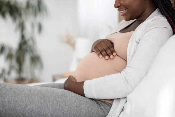 Mulher grávida preta sentada na cama abraçando a barriga, vista lateral — Fotografia de Stock