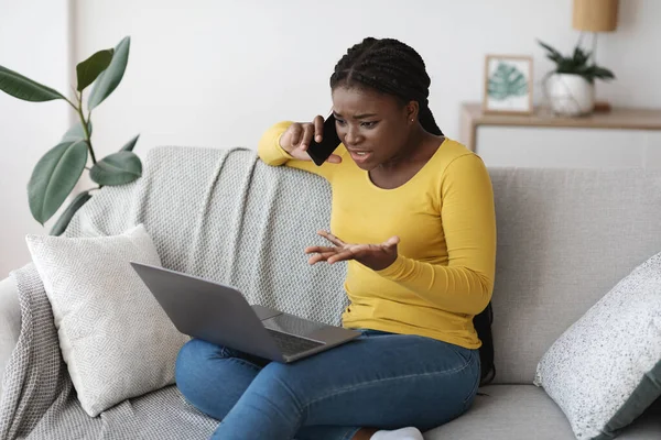 Frustrated black lady using laptop and talking on cellphone at home — Stock Photo, Image