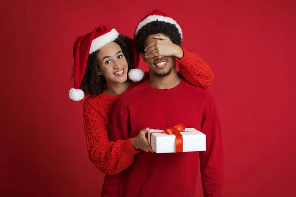 Sonriente joven afroamericana millennial mujer en Santa Claus sombrero cierra los ojos del hombre en suéter y le da regalo — Foto de Stock