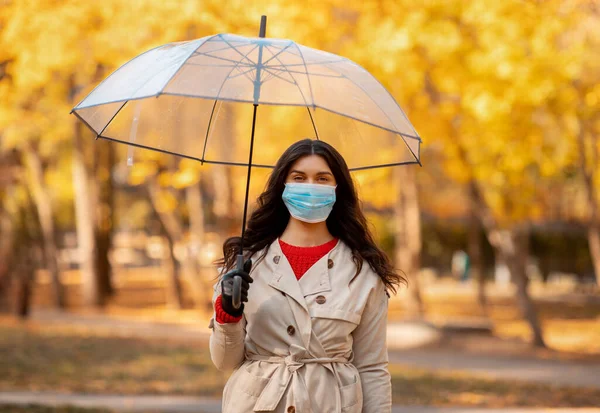 Mooie millenial dame met paraplu en medisch masker staan onder de regen en kijken naar de camera in de herfst park — Stockfoto