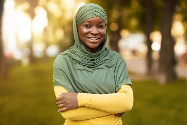 Smiling Black Muslim Woman In Hijab Standing With Folded Arms Outdoors