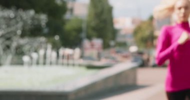 Genç sporcu kadın şehir parkında koşuyor, çeşmenin yanında nefes almak için duruyor ve koşmaya devam ediyor, ağır çekim.