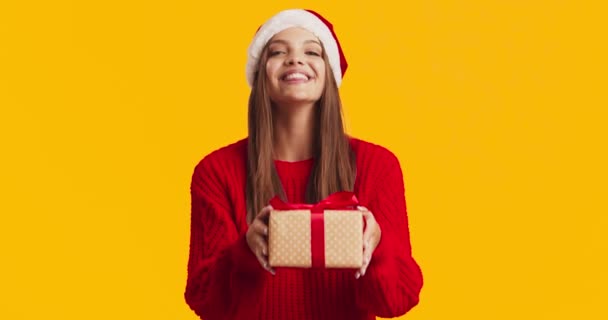 Entzückende junge Dame mit Weihnachtsmütze und rotem Pullover bietet Geschenkbox vor der Kamera und lächelnden, orangefarbenen Studiohintergrund — Stockvideo