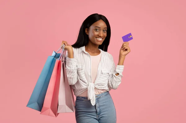 Mooie zwarte vrouw met credit card houden geschenkzakjes en glimlachen op roze studio achtergrond — Stockfoto
