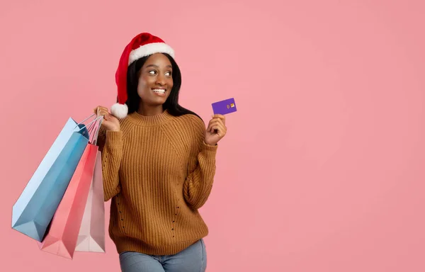 Les courses de Noël. Jolie femme noire dans le chapeau Santa tenant des sacs-cadeaux et carte de crédit sur fond de studio rose, espace de copie — Photo