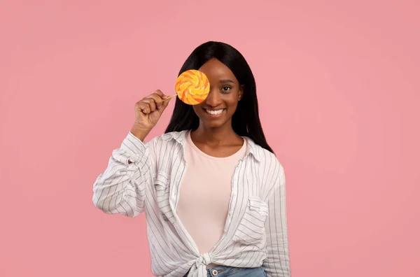 Smiling African American woman in casual clothes holding bright lollipop on pink studio background — Stock Photo, Image