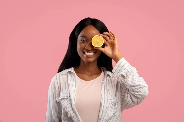 Vitamina C para o conceito de cuidados com a pele. Mulher negra bonita fechando o olho com limão metade no fundo do estúdio rosa — Fotografia de Stock