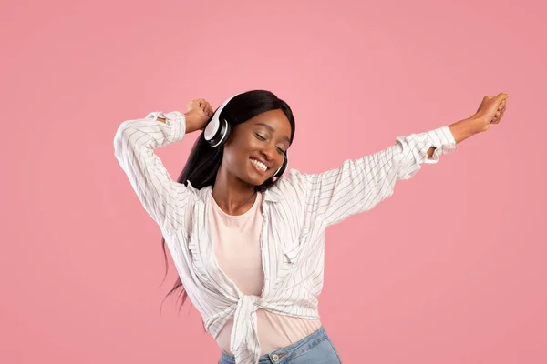 Mulher negra feliz no fone de ouvido dançando a bela música no fundo do estúdio rosa — Fotografia de Stock