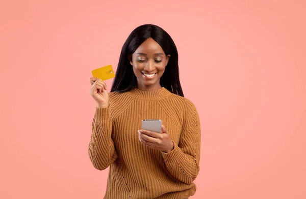 Інтернет-магазин. Позитивна чорна жінка з мобільним телефоном та кредитною карткою купує речі через рожеве тло. — стокове фото