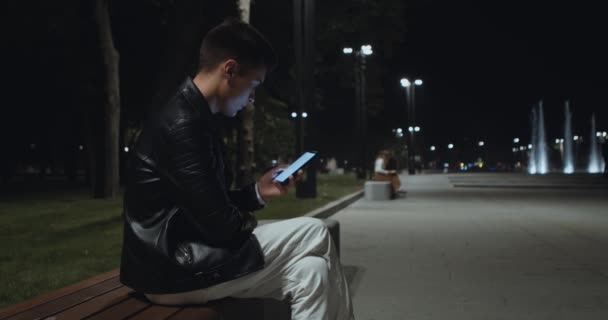 Junger Mann netzt am Handy, sitzt allein auf Bank im Abendpark — Stockvideo