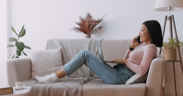 Pozitív ázsiai hölgy szabadúszó beszél telefonon ügyfél és dolgozik laptop, fekvő kanapén otthon, oldalnézet — Stock videók
