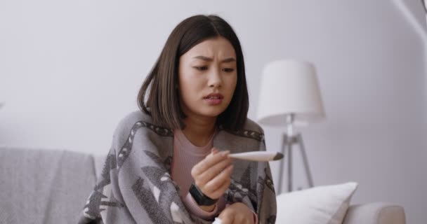 Ideges ázsiai nő, aki lázas lázmérőt néz, aggódik a koronavírus tünetei miatt, lelassult a mozgása. — Stock videók
