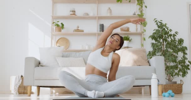 Yoga en fitness. Jonge zwarte dame zitten in lotus positie en strekken haar lichaam aan de zijkanten thuis, slow motion — Stockvideo