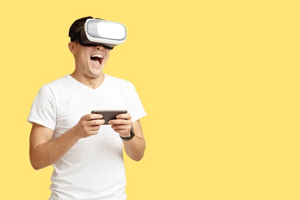 Happy young man with open mouth in virtual reality glasses holds smartphone and play — Stock Photo, Image