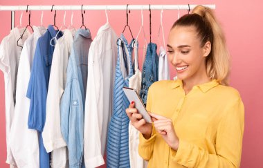 Gülümseyen kadın elinde telefonla rafın yanında duruyor.