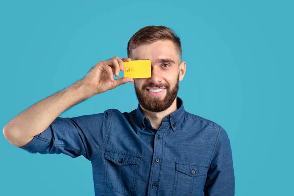 Sorrindo millennial cara segurando cartão de crédito na frente de seu olho no fundo do estúdio azul — Fotografia de Stock