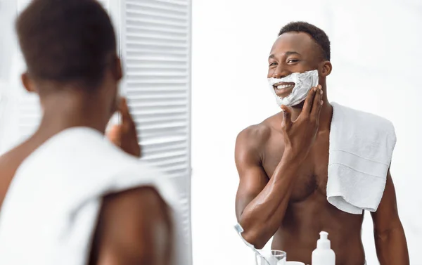 Africano chico aplicando afeitar espuma preparándose para afeitarse en baño — Foto de Stock