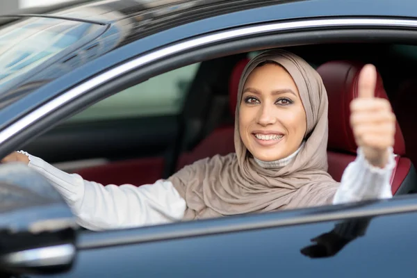Positive muslim woman driving car, showing thumbs up