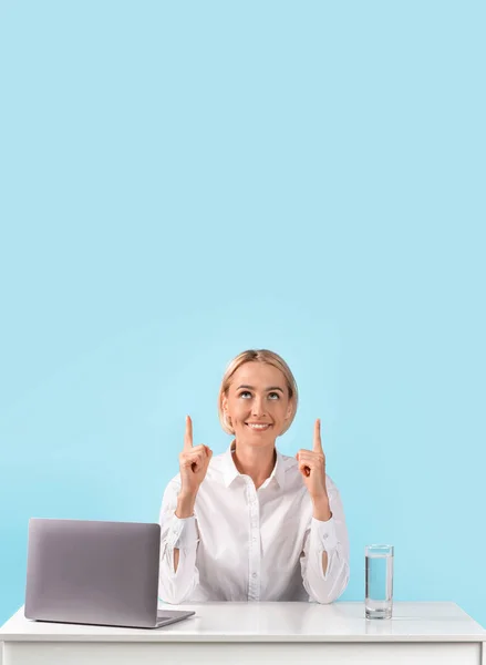 Щаслива бізнес-леді сидить за столом з ноутбуком і вказує вгору на порожнє місце, синій студійний фон — стокове фото