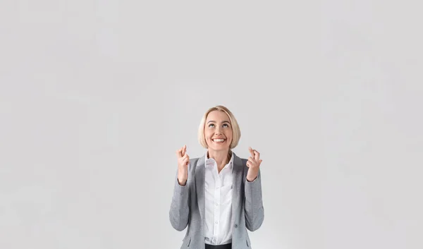 Felice donna d'affari bionda in abito formale incrociando le dita per buona fortuna su sfondo grigio chiaro studio, spazio copia — Foto Stock