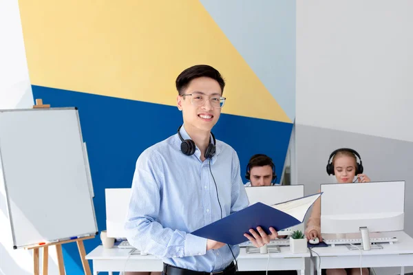 Operador de helpdesk masculino asiático feliz em headset segurando documentos no escritório moderno — Fotografia de Stock