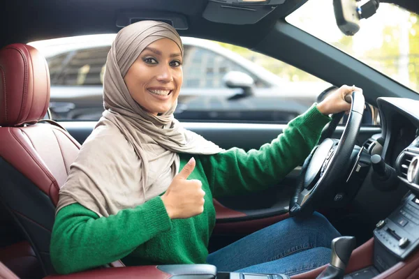 Cheerful muslim woman driving car, showing thumbs up