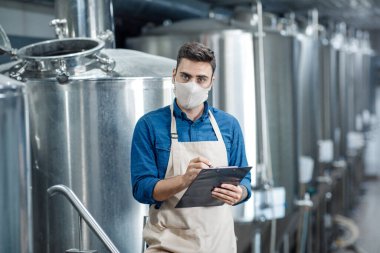 Ciddi bir adam karantina sırasında bira fabrikasında duruyor ve ekipman verilerini yazıyor.