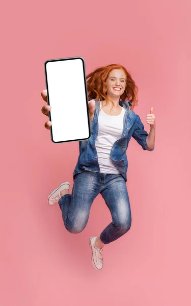 Happy redhead girl jumping with smartphone and showing thumb up — Stock Photo, Image