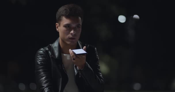 Розпізнавання голосу. Молодий серйозний чоловік записує звукове повідомлення на мобільний телефон, сидячи в міському парку пізно ввечері — стокове відео