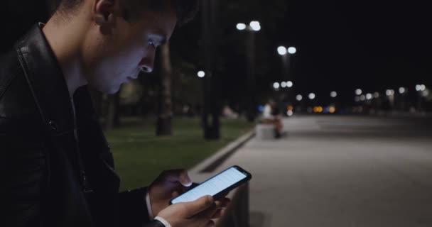 Young lonely guy sitting alone in evening park and networking on smartphone, close up — Stock Video