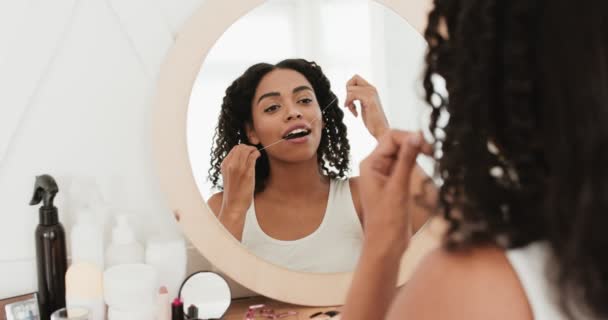 Mujer afroamericana joven sentada frente al espejo y usando hilo dental para cepillarse los dientes en el baño, vista trasera — Vídeos de Stock