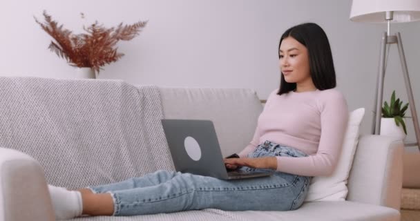 Online kommunikáció. Boldog nyugodt fiatal ázsiai nő ül a kanapén, és gépel a laptopon