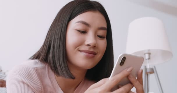 Nahaufnahme Porträt einer fröhlichen asiatischen Frau, die auf ihrem Handy SMS schreibt und mit Freunden zu Hause chatten kann — Stockvideo