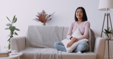 Asyalı güzellik. Evdeki koltukta oturan genç mutlu Çinli kadın kameraya gülüyor, yastığı kucaklıyor.
