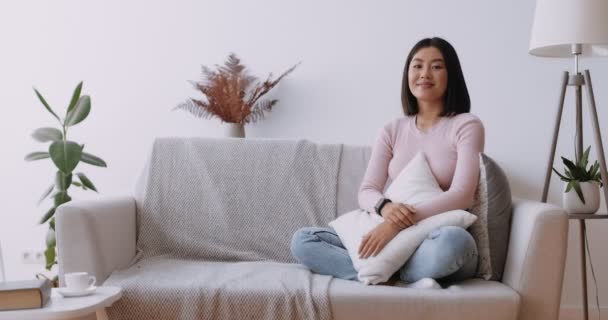 Bellezza asiatica. Giovane signora cinese felice seduta sul divano a casa e ridendo della macchina fotografica, abbracciando cuscino — Video Stock