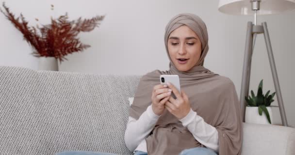 Jeune femme musulmane surprise dans le hijab en utilisant un smartphone à la maison, messagerie avec des amis, assis sur le canapé dans le salon — Video