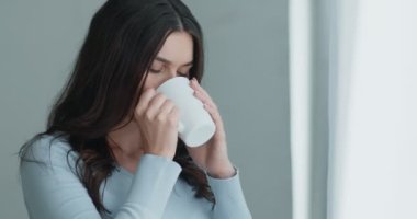 Endişeli genç bir kadın, pencereden dışarı bakıyor, elinde bir fincan sıcak çay, ağır çekimde