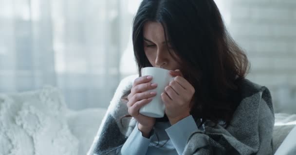 Joven mujer deprimida calentando las manos de la taza, disfrutando del té caliente, sentado en el sofá solo envuelto en cuadros — Vídeo de stock