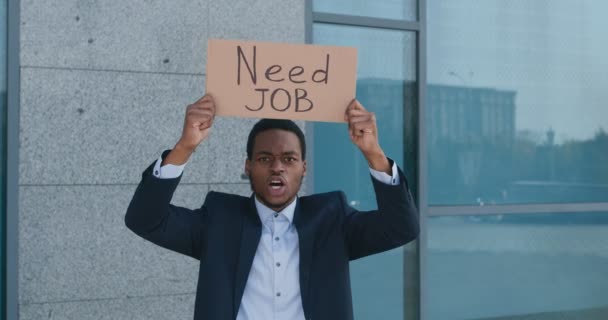 Отчаянный африканский американец в формальном костюме с плакатом NEED JOB и кричащий возле офисного здания, следящий выстрел — стоковое видео