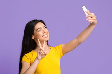 Selfie Eğlencesi. Neşeli Asyalı kadın akıllı telefonda kendi portresini çekiyor, barış jestleri gösteriyor.