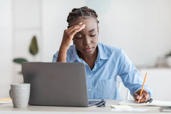 Cansada empleada de oficina negra sentada en el escritorio con laptop, tomando notas — Foto de Stock