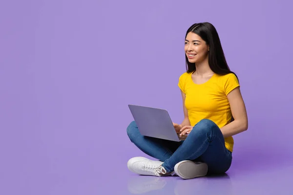 Uśmiechnięta azjatka siedzi na podłodze z laptopem na fioletowym tle — Zdjęcie stockowe