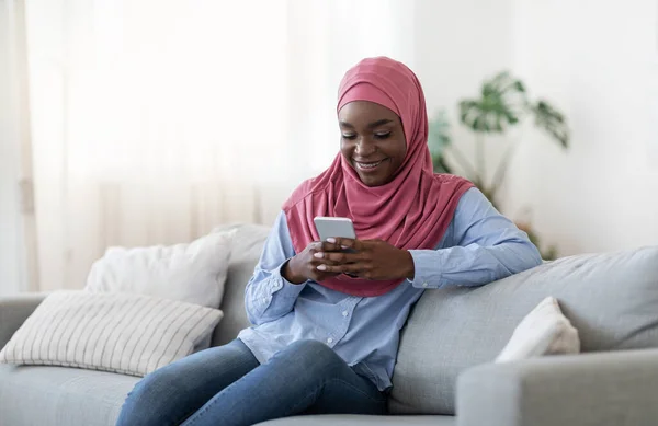 Χαλαρωμένη μαύρη μουσουλμάνα γυναίκα κάθεται στον καναπέ Μηνύματα στο Smartphone στο σπίτι — Φωτογραφία Αρχείου
