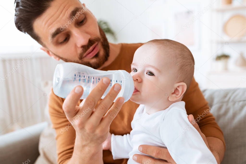 Dad Giving Bottle To Baby Sitting On Couch At Home