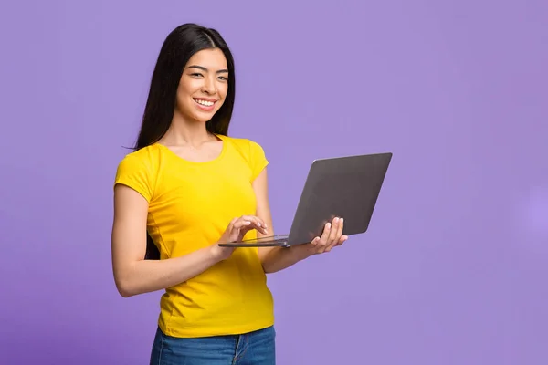 Pozytywne młoda Azjatka pozowanie z laptopa komputera na fioletowym tle — Zdjęcie stockowe
