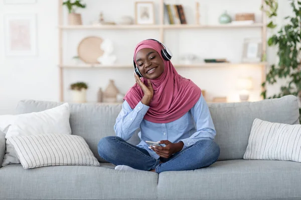 Black Muslim Woman Wearing Wireless Headphone, Listening Music On Smartphone At Home — Stok Foto